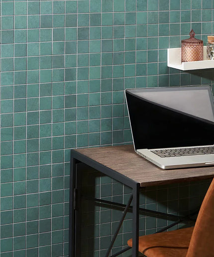 Laptop on a wooden desk against a teal tiled wall with a shelf holding decorative items.
