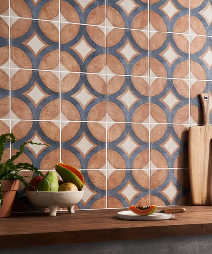 Patterned tiles on a wall with a wooden shelf below containing fruits, a plant, and kitchen utensils.