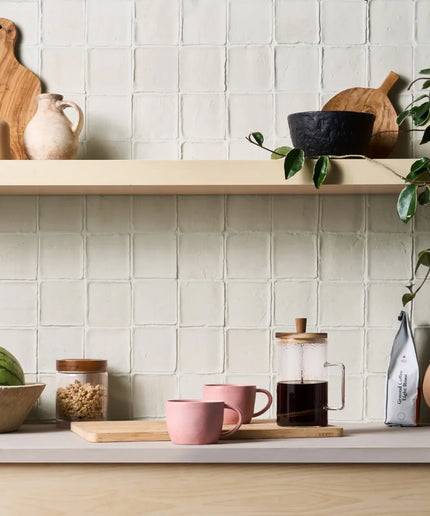 Kitchen counter with coffee setup, mugs, and a plant against a tiled wall.