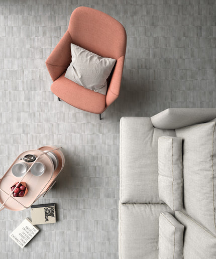 A top-down view of a modern living room corner featuring a dusty pink armchair with a light gray pillow, a light gray sofa, and a matching pink side table holding a tray with cups and a small decorative object, all set on a gray textured tile floor.
