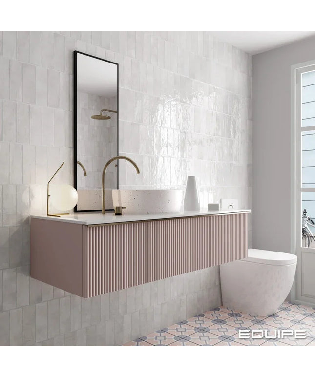 A modern floating bathroom vanity in a dusty pink ribbed finish, paired with a white vessel sink, a white marble countertop, and a tall black-framed mirror. The bathroom features textured light gray tiles, a white wall, and a patterned tile floor, with a window on the right.