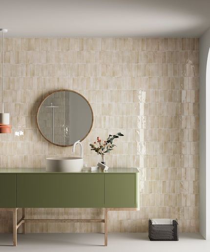 A modern bathroom vanity setup featuring glossy beige rectangular wall tiles, a sage green cabinet with a white vessel sink, a round wood-framed mirror, and a hanging pendant light.