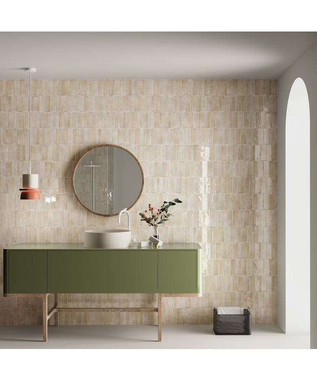 A modern bathroom vanity setup featuring glossy beige rectangular wall tiles, a sage green cabinet with a white vessel sink, a round wood-framed mirror, and a hanging pendant light.