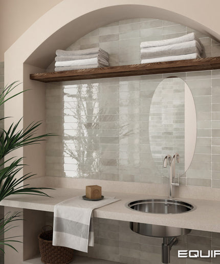 A bathroom vanity area with a light stone countertop, round stainless steel sink, oval mirror, and pale green subway tile backsplash. A wooden shelf above holds folded white towels, and a potted plant sits at the left edge.
