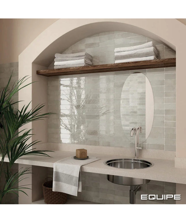 A bathroom vanity area with a light stone countertop, round stainless steel sink, oval mirror, and pale green subway tile backsplash. A wooden shelf above holds folded white towels, and a potted plant sits at the left edge.