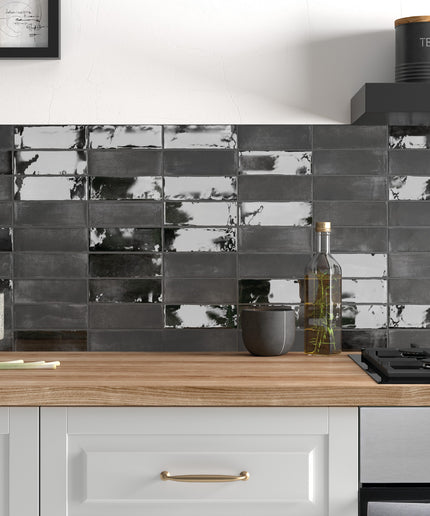 A modern kitchen backsplash featuring glossy rectangular black and gray tiles with a reflective, marbled finish, mounted above a light wood countertop with white cabinets.