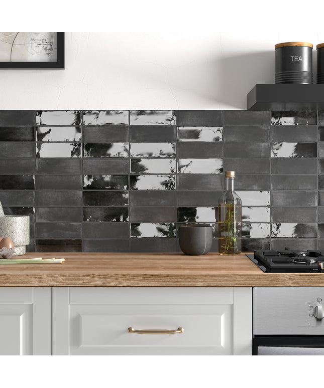 A modern kitchen backsplash featuring glossy rectangular black and gray tiles with a reflective, marbled finish, mounted above a light wood countertop with white cabinets.