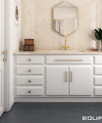 A white bathroom vanity cabinet with gold hardware, a marble countertop, and a gold faucet, shown in a styled bathroom setting with a beige herringbone tile backsplash, a hexagonal mirror, and a dark gray floor. The brand name 'EQUIPE' is visible in the bottom right corner.