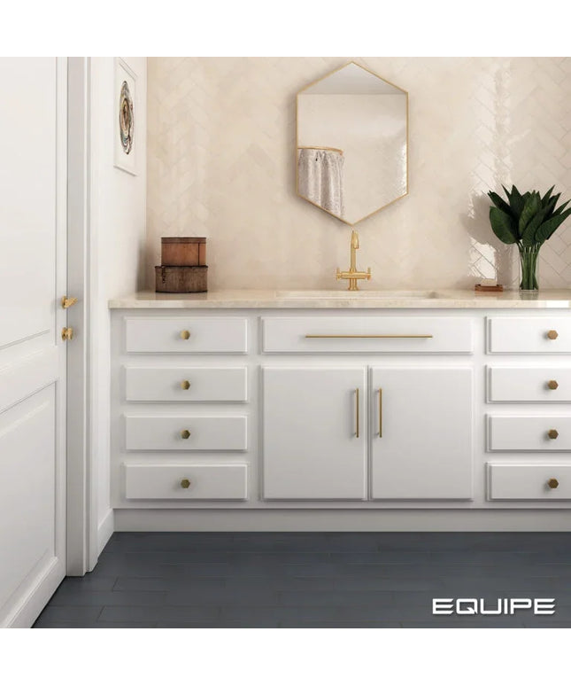 A white bathroom vanity cabinet with gold hardware, a marble countertop, and a gold faucet, shown in a styled bathroom setting with a beige herringbone tile backsplash, a hexagonal mirror, and a dark gray floor. The brand name 'EQUIPE' is visible in the bottom right corner.