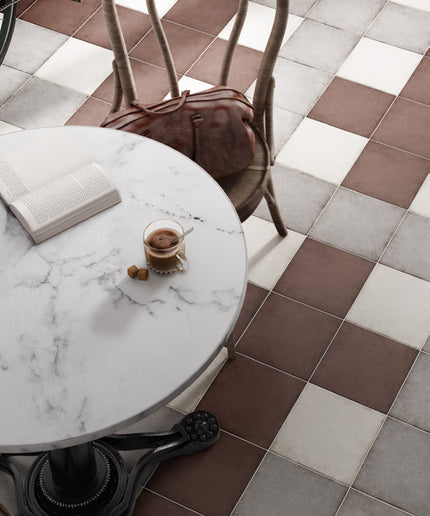 Brown, white, and gray checkered floor tiles in a cafe setting, with a round white marble table, a wooden chair with a brown leather bag, and a glass of coffee.