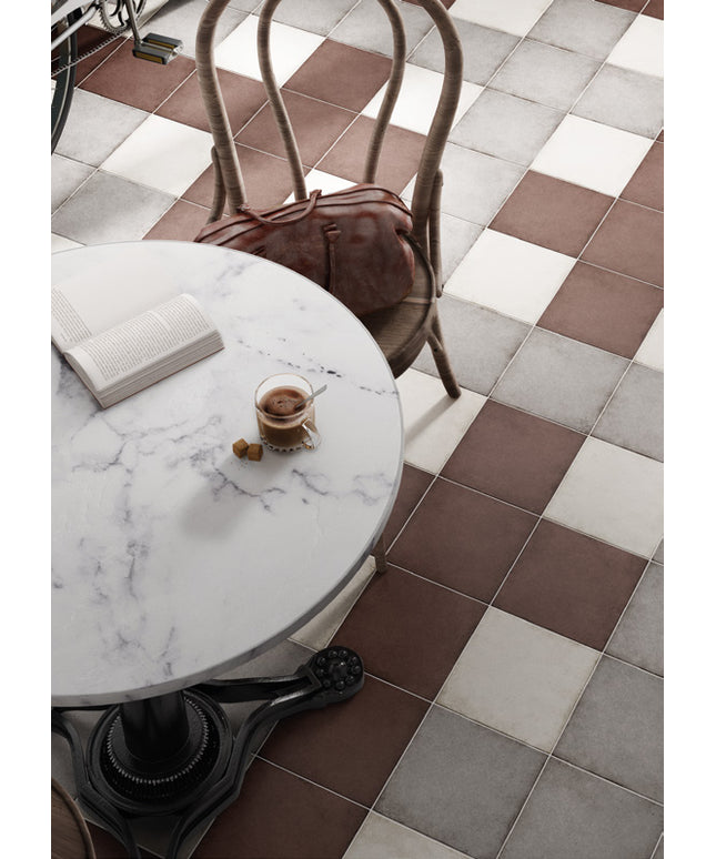 Brown, white, and gray checkered floor tiles in a cafe setting, with a round white marble table, a wooden chair with a brown leather bag, and a glass of coffee.