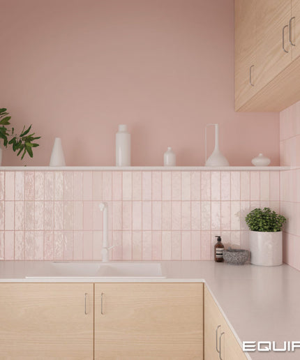 A modern kitchen corner with light wood cabinets, a white countertop, and a pink tiled backsplash. White ceramic vases and a small potted plant sit on a shelf against a soft pink wall.