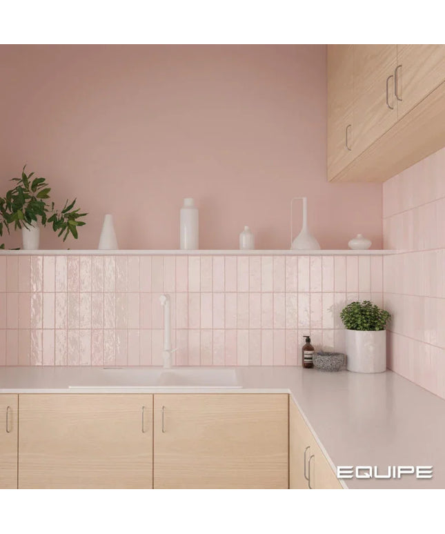 A modern kitchen corner with light wood cabinets, a white countertop, and a pink tiled backsplash. White ceramic vases and a small potted plant sit on a shelf against a soft pink wall.