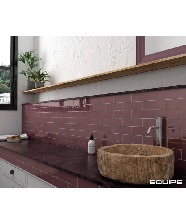 A bathroom vanity setup featuring glossy terracotta pink subway tiles, a dark stone countertop, a round natural wood vessel sink, and a white wall with a wooden shelf and plant.