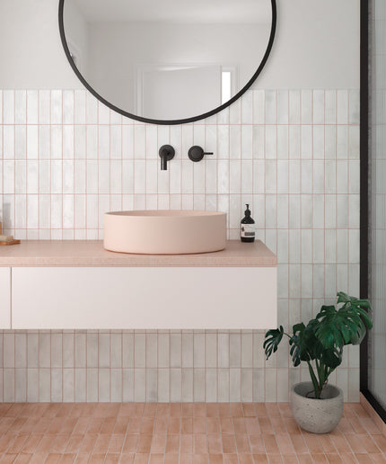 A modern bathroom vanity setup with a round blush pink vessel sink on a light stone countertop, set against a white subway tile wall with a black-framed round mirror above. A potted green plant sits on the terracotta tile floor next to a glass shower door.