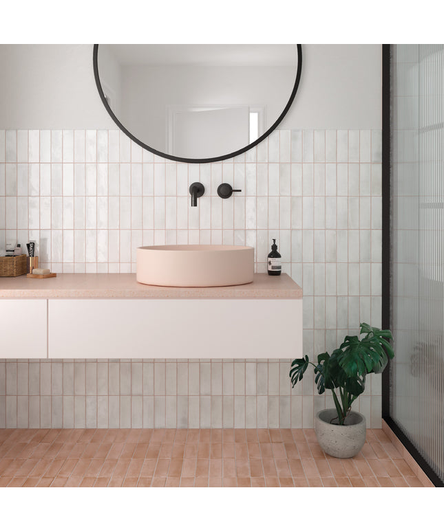 A modern bathroom vanity setup with a round blush pink vessel sink on a light stone countertop, set against a white subway tile wall with a black-framed round mirror above. A potted green plant sits on the terracotta tile floor next to a glass shower door.