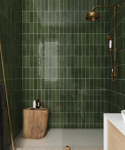 A modern shower area with glossy dark green rectangular wall tiles, a brass shower head and hand shower mounted on the wall, a glass door, a small wooden stool, and part of a white vanity visible in the foreground.
