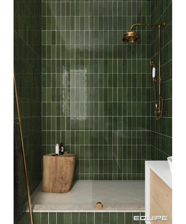 A modern shower area with glossy dark green rectangular wall tiles, a brass shower head and hand shower mounted on the wall, a glass door, a small wooden stool, and part of a white vanity visible in the foreground.