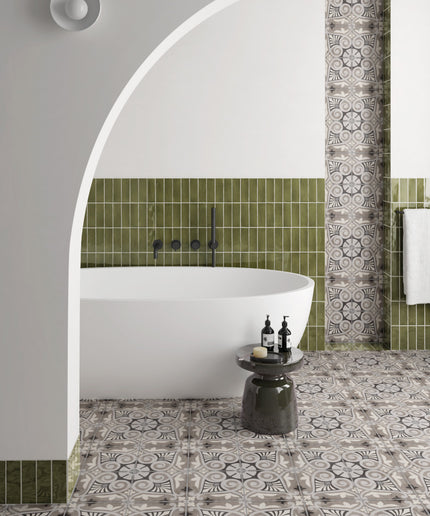 A styled bathroom interior featuring a white freestanding bathtub set within a white arched alcove, surrounded by olive green square tiles and patterned gray-and-white floor and wall tiles.