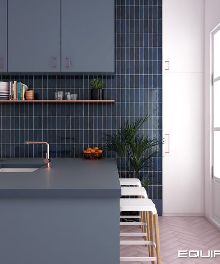A modern kitchen interior with a blue-gray island, dark blue subway tile backsplash, copper-toned faucet, white bar stools, and a large window on the right. The word EQUIPE appears in the bottom right corner.
