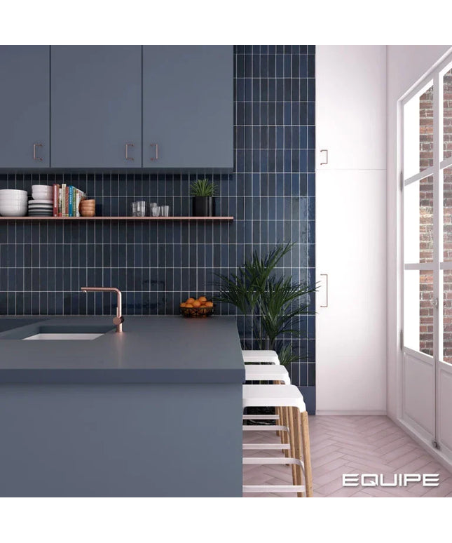 A modern kitchen interior with a blue-gray island, dark blue subway tile backsplash, copper-toned faucet, white bar stools, and a large window on the right. The word EQUIPE appears in the bottom right corner.