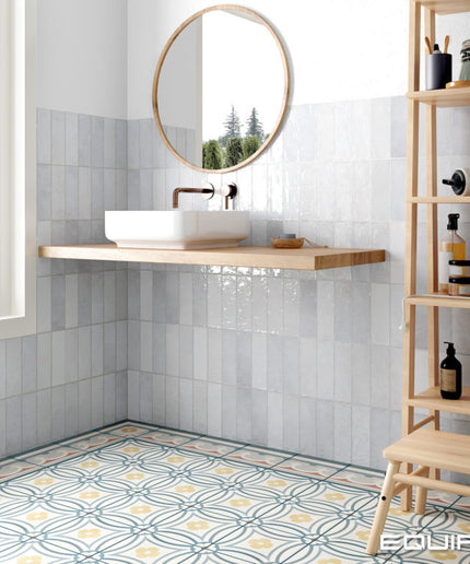 A modern bathroom corner with glossy white wall tiles, a floating wooden sink shelf, a white vessel sink, a round wood-framed mirror, patterned blue and yellow floor tiles, and a tall light wood shelving unit on the right.
