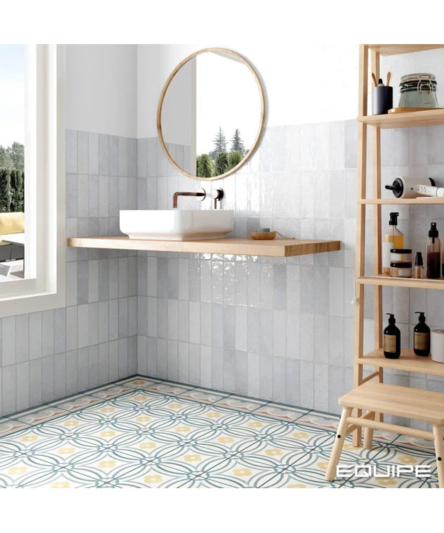 A modern bathroom corner with glossy white wall tiles, a floating wooden sink shelf, a white vessel sink, a round wood-framed mirror, patterned blue and yellow floor tiles, and a tall light wood shelving unit on the right.