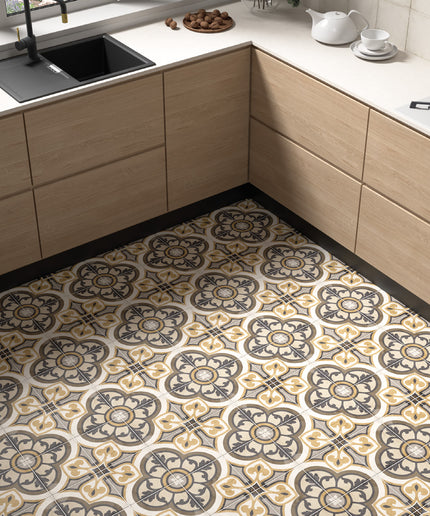 A kitchen interior showing a patterned floor tile with a repeating ornate medallion design in beige, gray, and black, installed in a modern L-shaped kitchen with light wood cabinets and white countertops.