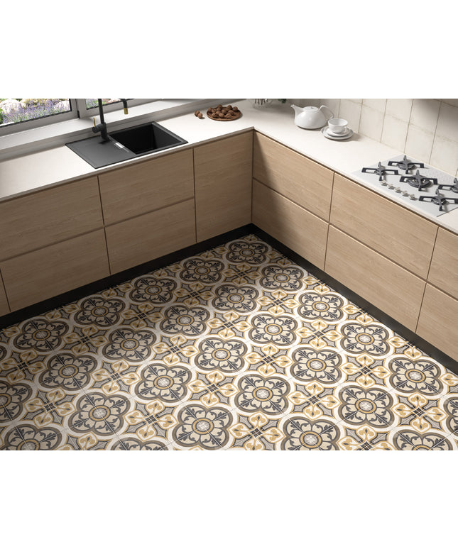 A kitchen interior showing a patterned floor tile with a repeating ornate medallion design in beige, gray, and black, installed in a modern L-shaped kitchen with light wood cabinets and white countertops.