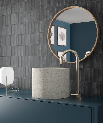 A modern bathroom vanity setup featuring a cylindrical speckled stone sink, a curved matte brass faucet, a round wood-framed mirror, and a small white lamp, all set against a dark gray tiled wall and a deep blue vanity cabinet.