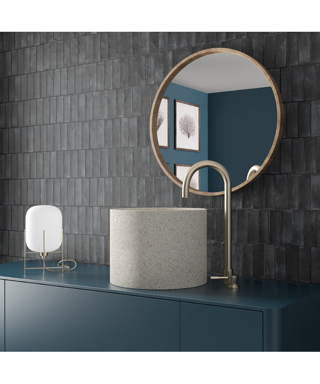 A modern bathroom vanity setup featuring a cylindrical speckled stone sink, a curved matte brass faucet, a round wood-framed mirror, and a small white lamp, all set against a dark gray tiled wall and a deep blue vanity cabinet.