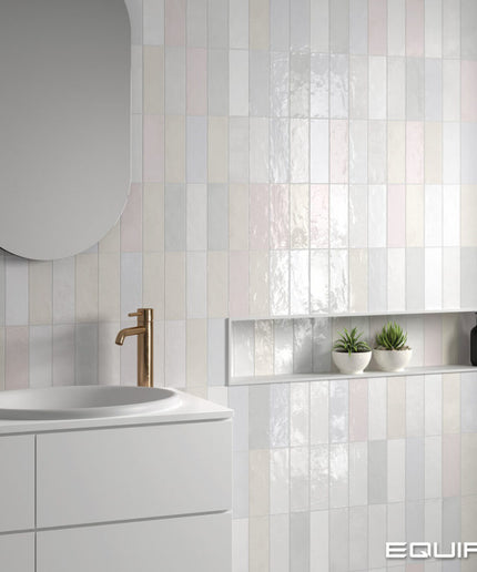 A modern bathroom interior featuring glossy rectangular wall tiles in soft vertical stripes of white, pale pink, and light gray. A white vanity with a white sink and a bronze faucet is visible on the left, and a recessed shelf with two small potted succulents and a black bottle is mounted on the right.