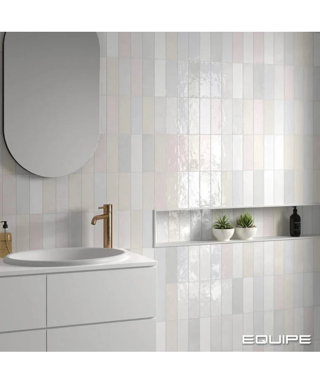 A modern bathroom interior featuring glossy rectangular wall tiles in soft vertical stripes of white, pale pink, and light gray. A white vanity with a white sink and a bronze faucet is visible on the left, and a recessed shelf with two small potted succulents and a black bottle is mounted on the right.