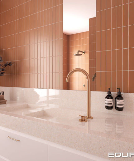 A modern bathroom vanity setup with a matte copper gooseneck faucet mounted on a white speckled countertop. The background features a large mirror reflecting a tiled wall in a warm terracotta orange-brown color, with white cabinets below.