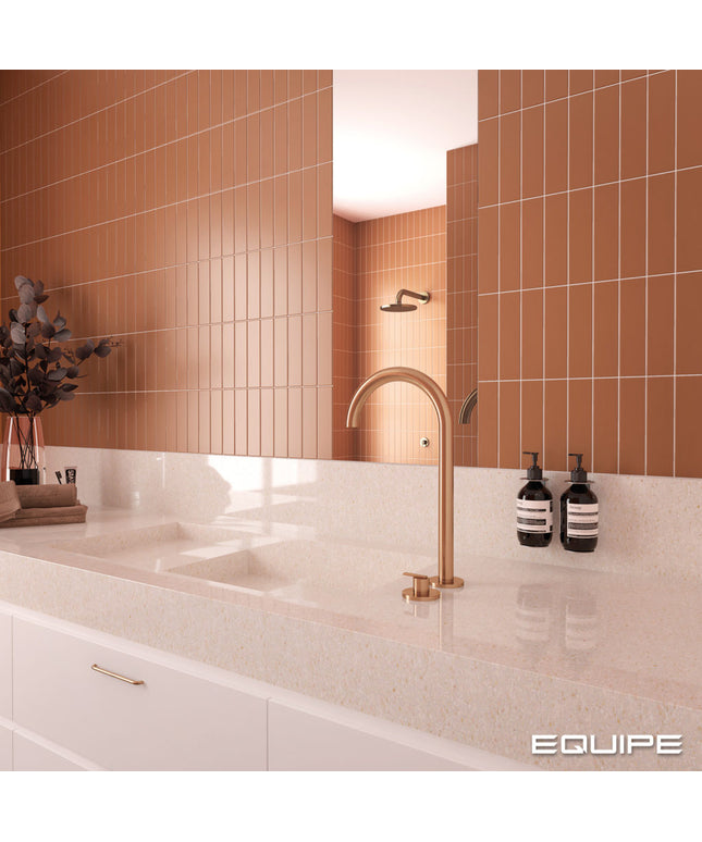 A modern bathroom vanity setup with a matte copper gooseneck faucet mounted on a white speckled countertop. The background features a large mirror reflecting a tiled wall in a warm terracotta orange-brown color, with white cabinets below.