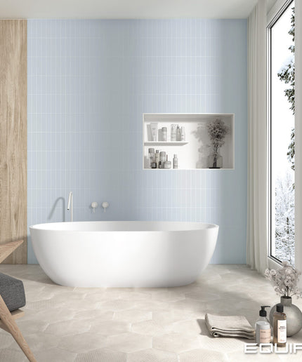 A modern freestanding oval bathtub in a bright bathroom with pale blue wall tiles, a recessed shelf with toiletries, a wood cabinet, and a large window looking out onto a snowy outdoor scene. The brand name EGUIPE appears in the bottom right corner.
