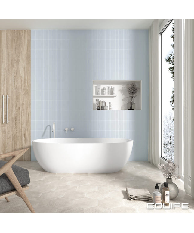 A modern freestanding oval bathtub in a bright bathroom with pale blue wall tiles, a recessed shelf with toiletries, a wood cabinet, and a large window looking out onto a snowy outdoor scene. The brand name EGUIPE appears in the bottom right corner.