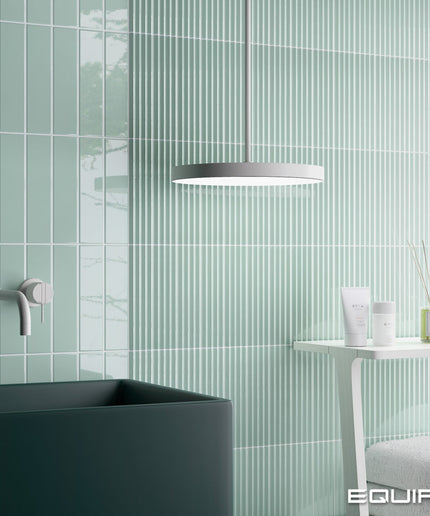 A modern bathroom scene with pale mint green vertically ribbed wall tiles, a dark charcoal sink, a white side table with toiletries, and a circular pendant light. The EQUITE brand name is visible in the bottom right corner.