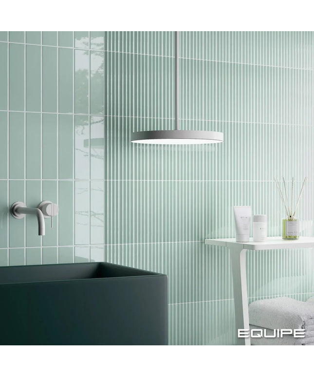 A modern bathroom scene with pale mint green vertically ribbed wall tiles, a dark charcoal sink, a white side table with toiletries, and a circular pendant light. The EQUITE brand name is visible in the bottom right corner.