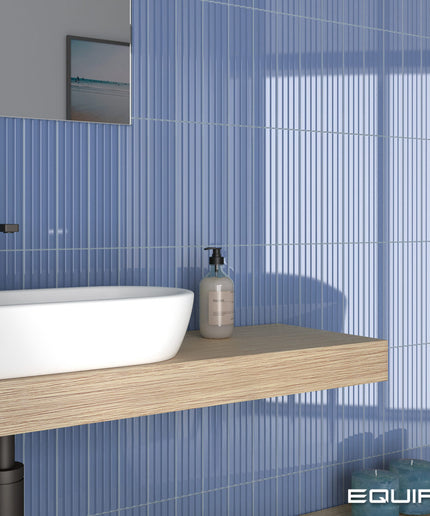 A modern bathroom scene featuring vertical blue ribbed wall tiles, a floating light wood vanity shelf, a white vessel sink, a black faucet, and a clear pump bottle. The brand name 'EQUIPE' is visible in the bottom right corner.