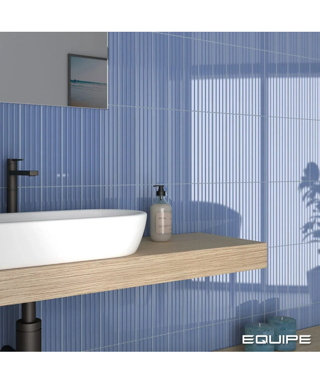 A modern bathroom scene featuring vertical blue ribbed wall tiles, a floating light wood vanity shelf, a white vessel sink, a black faucet, and a clear pump bottle. The brand name 'EQUIPE' is visible in the bottom right corner.