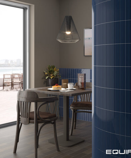 A modern dining area with a dark wood table and chairs, a black wire pendant light, and a prominent glossy blue ribbed cylindrical column on the right. A large window on the left looks out to a waterfront patio.