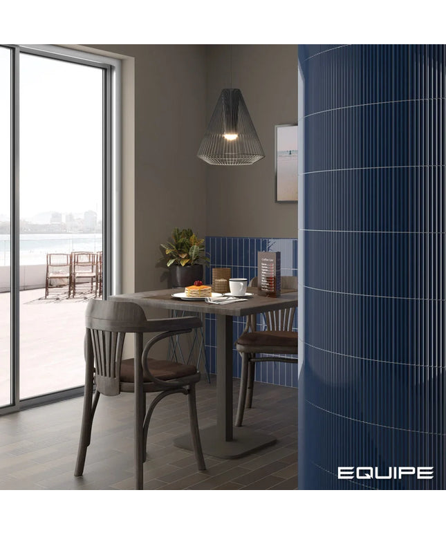 A modern dining area with a dark wood table and chairs, a black wire pendant light, and a prominent glossy blue ribbed cylindrical column on the right. A large window on the left looks out to a waterfront patio.