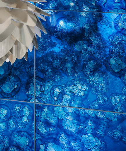 Blue patterned glass tiles with a bubbly, crystalline design, shown in a grid layout with dark grout lines. A white textured pendant light is partially visible in the top-left corner.