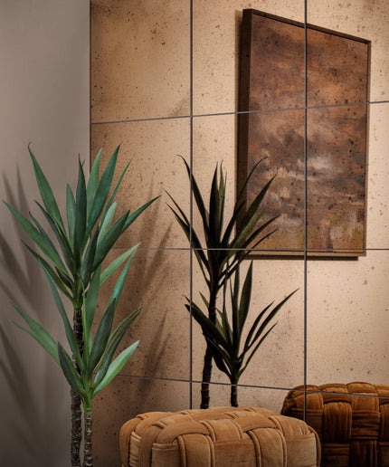 A styled interior corner with two potted green yucca plants, a large abstract framed artwork on a beige tiled wall, and two tufted upholstered ottomans in warm brown tones.