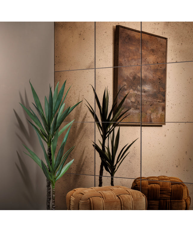 A styled interior corner with two potted green yucca plants, a large abstract framed artwork on a beige tiled wall, and two tufted upholstered ottomans in warm brown tones.