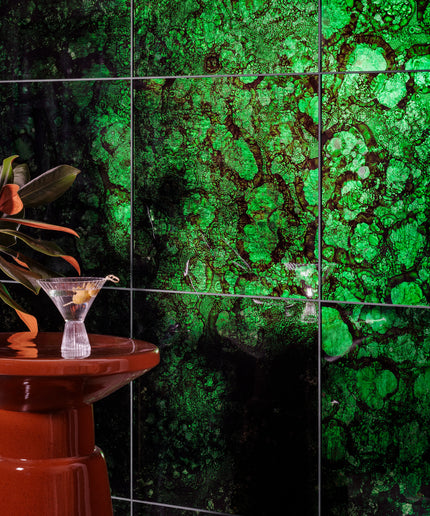 Green mottled stone-look ceramic wall tiles with dark grout lines, shown in a styled interior setting with a red pedestal and a cocktail glass.