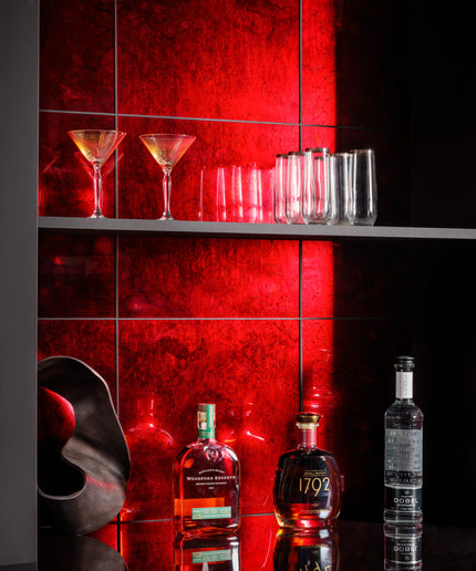 A styled bar scene with a red illuminated tiled wall, several clear drinking glasses on a shelf, and three liquor bottles on a reflective black counter below.