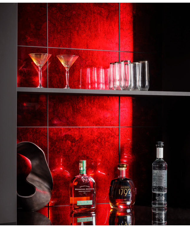 A styled bar scene with a red illuminated tiled wall, several clear drinking glasses on a shelf, and three liquor bottles on a reflective black counter below.