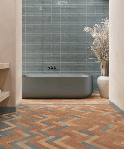 A modern bathroom interior with a freestanding matte gray bathtub, blue-gray rectangular wall tiles, terracotta and blue chevron floor tiles, beige walls, a built-in shelf, and a tall ceramic vase with dried grass.